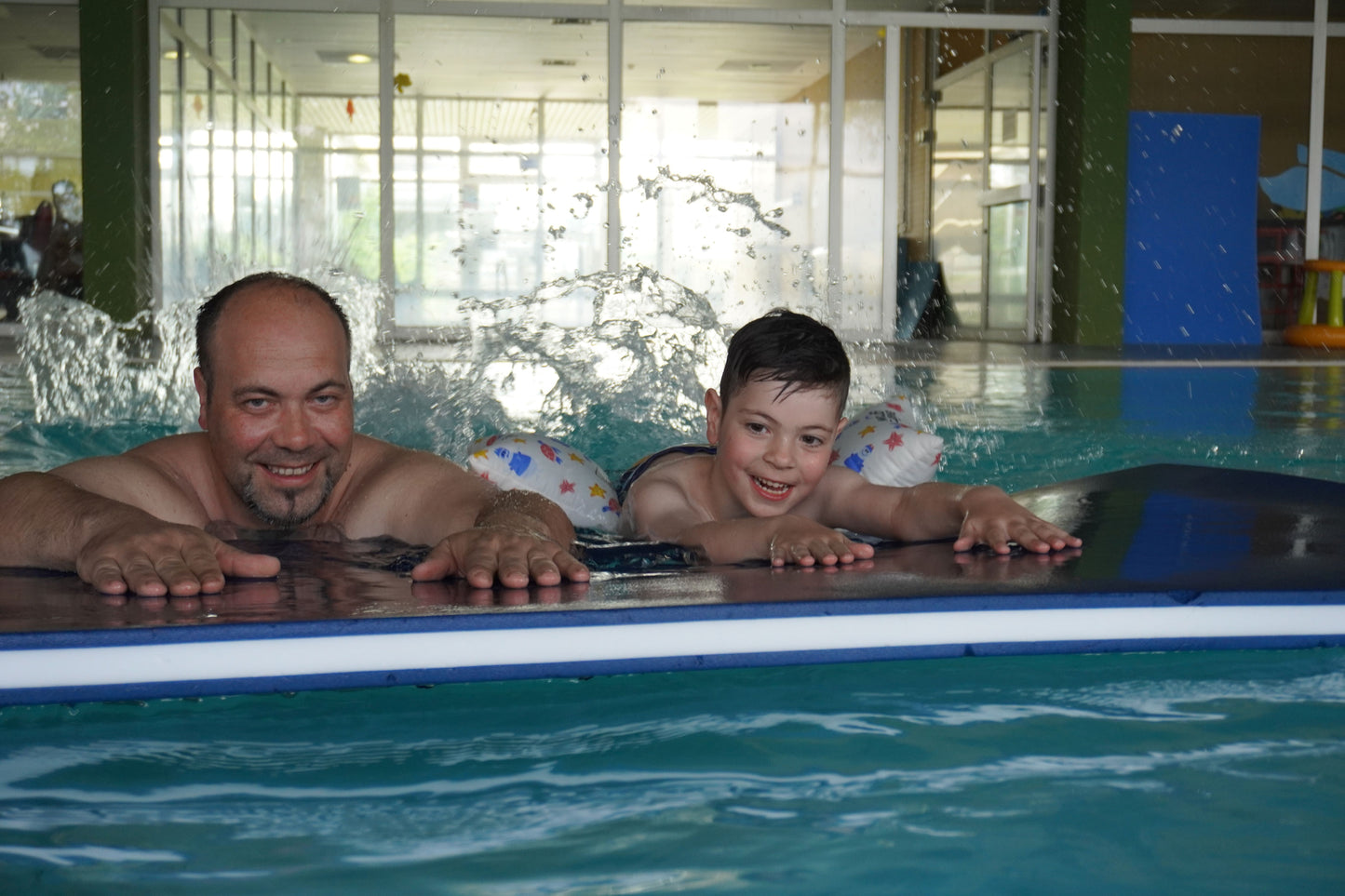 Vater und Sohn auf einer Matte, Kind hat Schwimmkissen an, beide lachen und strampeln im Wasser mit den Füßen