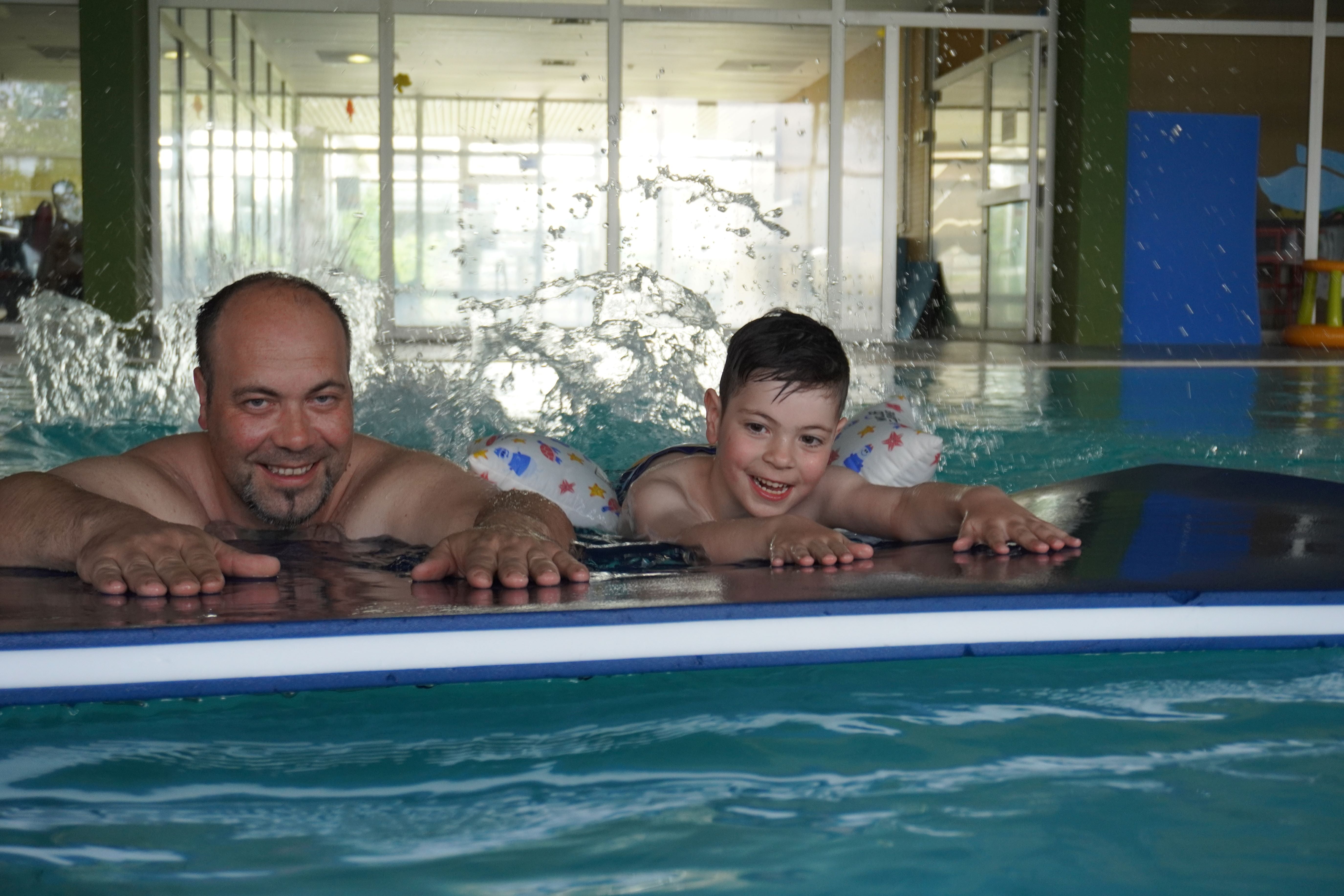 Vater und Sohn auf einer Matte, Kind hat Schwimmkissen an, beide lachen und strampeln im Wasser mit den Füßen